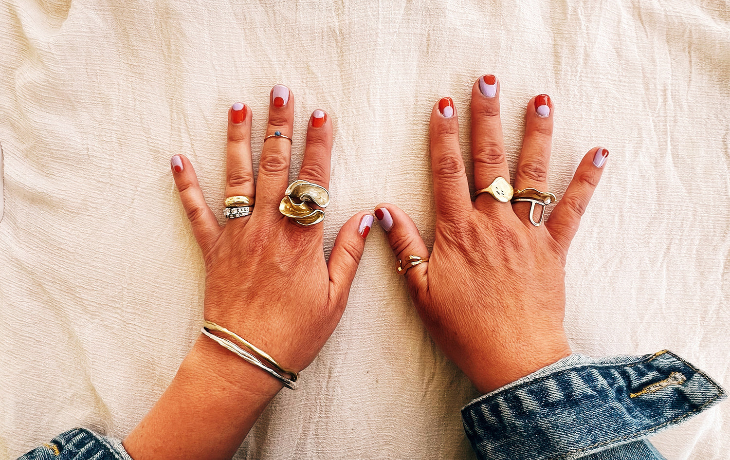two hands on a cream colored cloth wearing several rings and sporting colorful polish, and a couple of bangle bracelets
