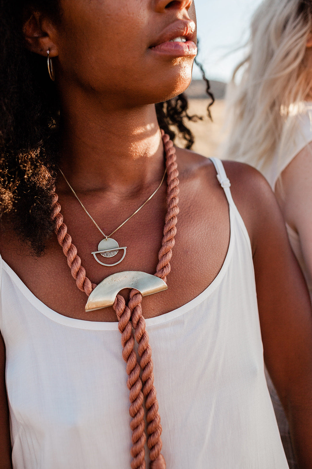 a dark skinned woman wearing two necklaces. One woven from twisted rosy thread with a large brass u shaped pendant.