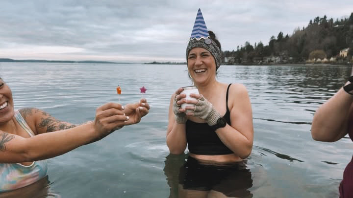 A woman wearing a birthday hat holding a cake while she stands in the Puget Sound cold plunging. She's surrounded by her friends who are holding birthay candles for her. 