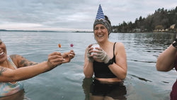 A woman wearing a birthday hat holding a cake while she stands in the Puget Sound cold plunging. She's surrounded by her friends who are holding birthay candles for her. 