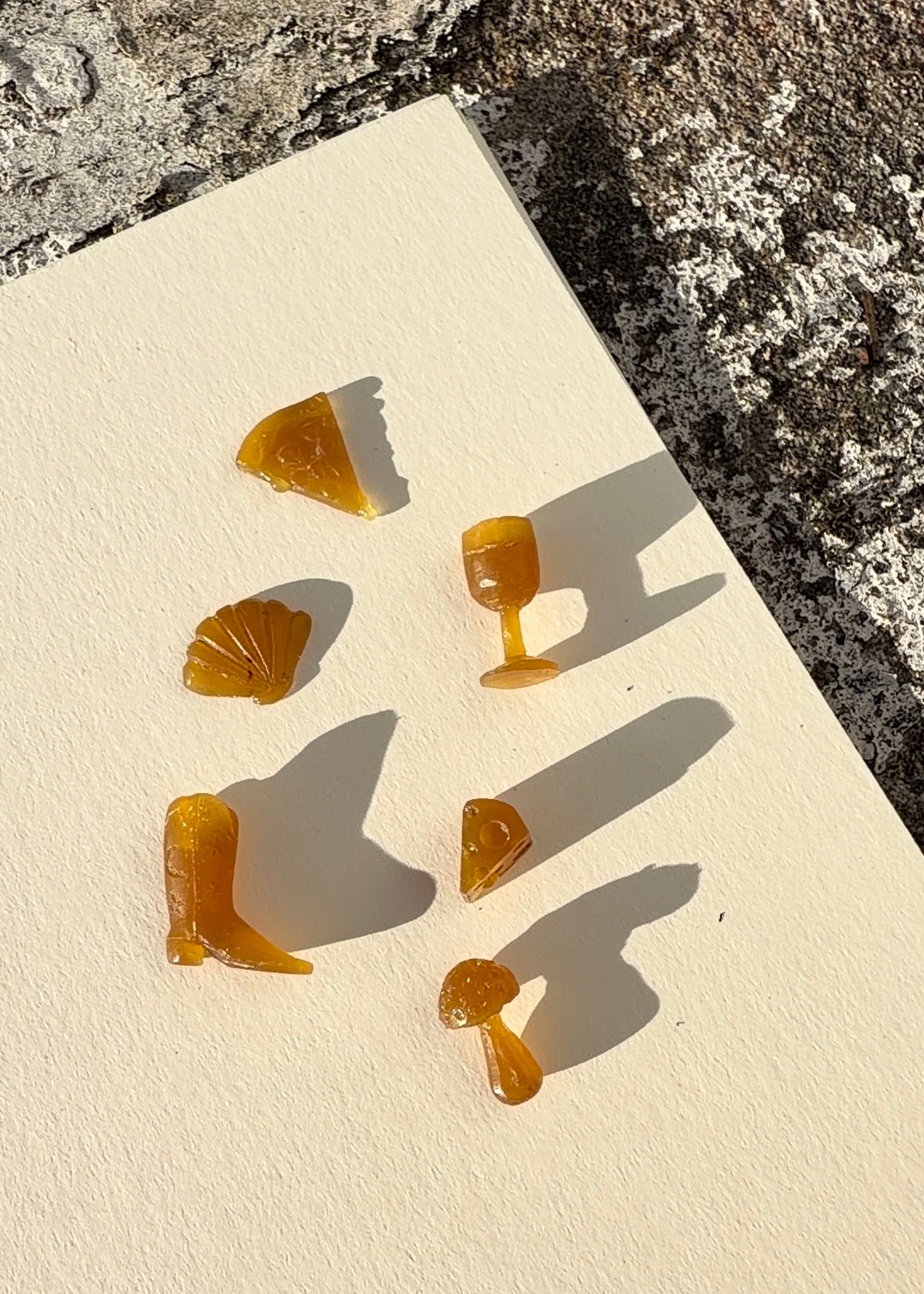 Six small wax carved charms arranged on a cream textured tile, with rough stone visible around the edges and strong sunlight casting sharp shadows.